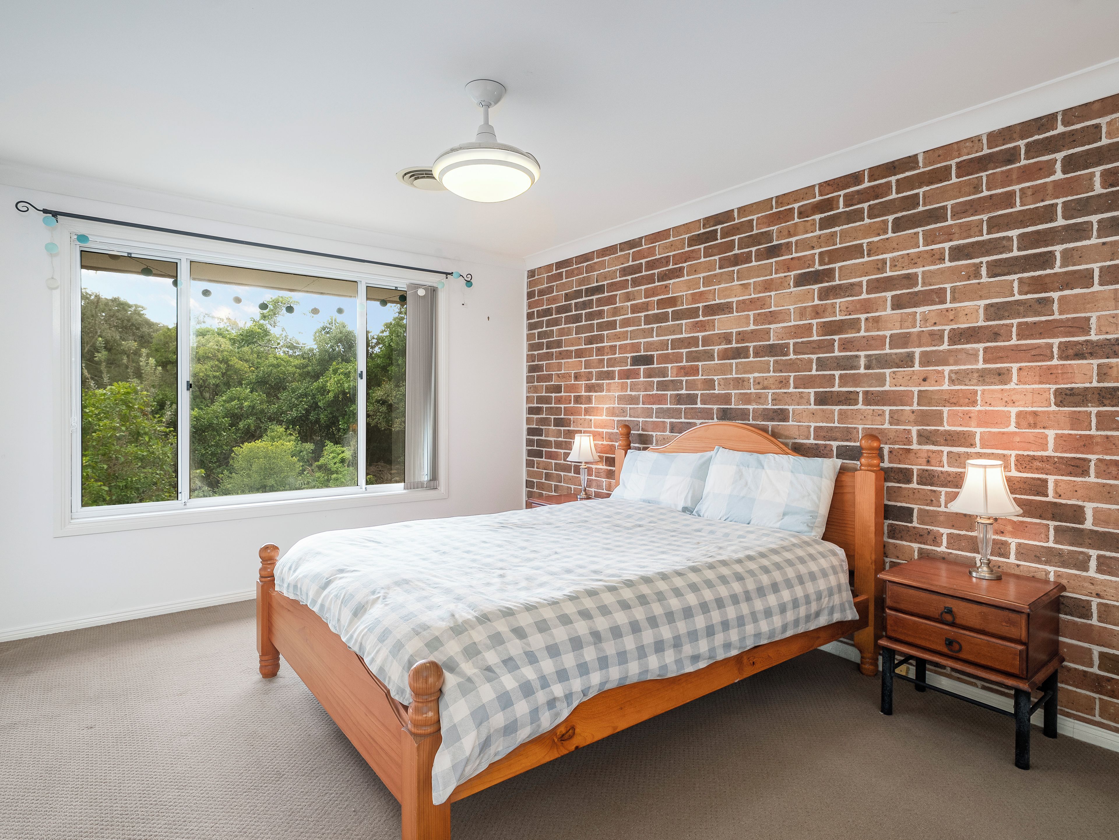 Une chambre avec un lit en bois, une table de chevet, une lampe, une fenêtre donnant sur un paysage verdoyant et un mur en briques.