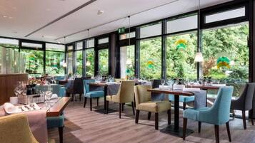 Ein Restaurant mit Tischen, Stühlen, großen Fenstern und Blick auf Grünflächen.