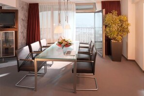 A modern hotel room with a glass dining table, leather chairs, a laptop, a decorative plant, a television, and a balcony with curtains.