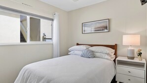 Un dormitorio con cama, mesita de noche, lámpara, un cuadro enmarcado y ventana con cortinas.