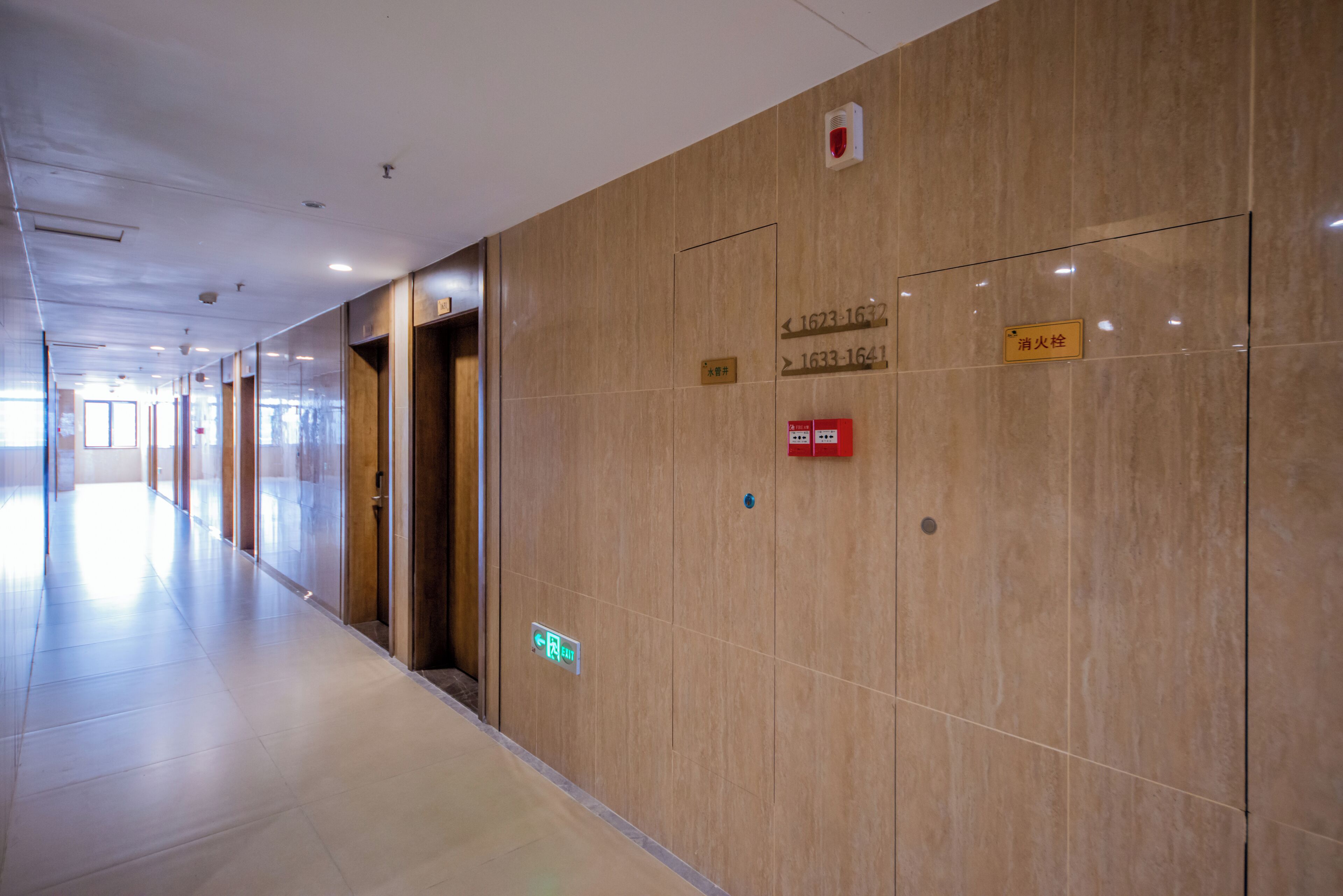 A long, well-lit corridor with marble walls, emergency exit signs, and fire safety equipment.