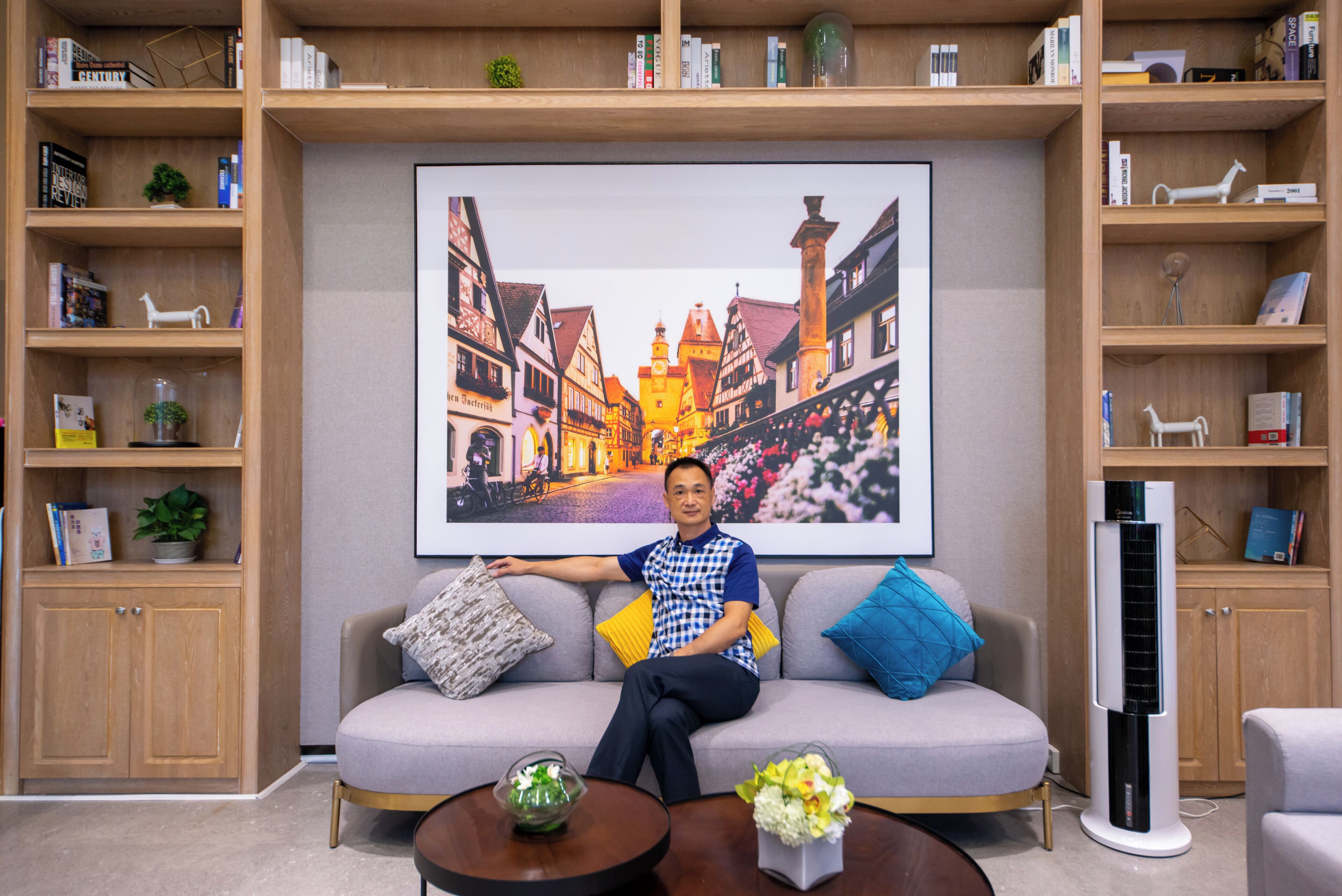 A man sitting on a sofa with a large framed picture of a European street behind him, surrounded by shelves with books and decorative items.