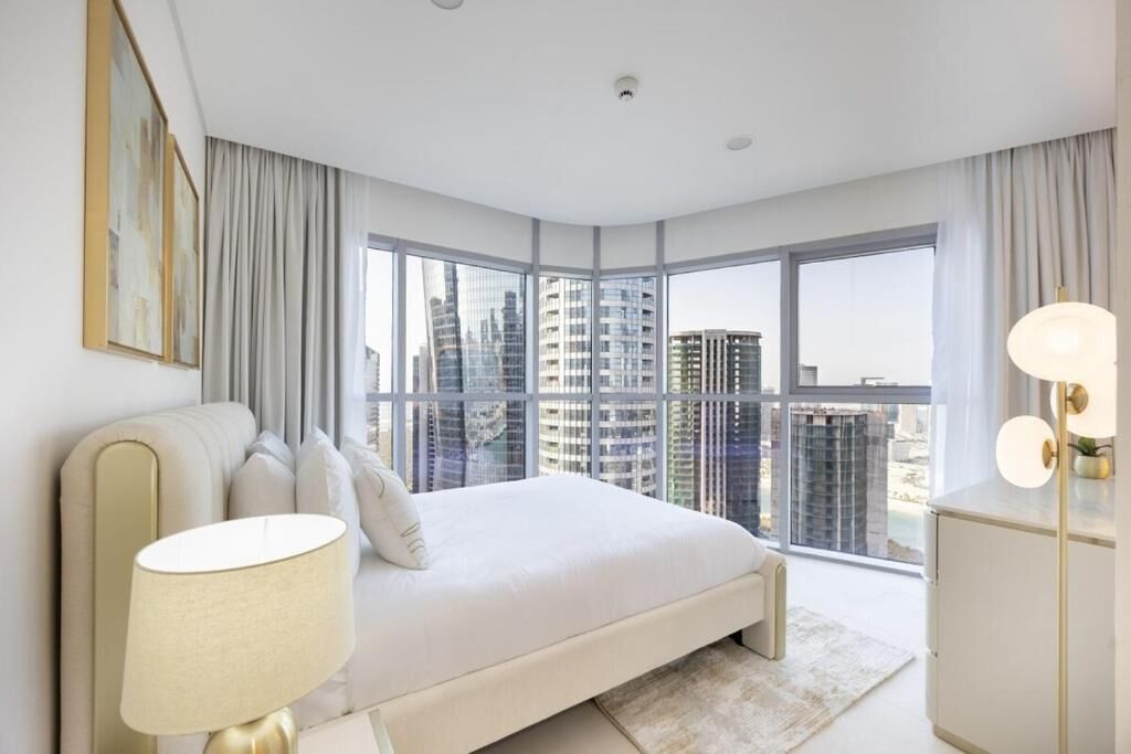 A modern bedroom with a large bed, a bedside lamp, and a view of high-rise buildings.