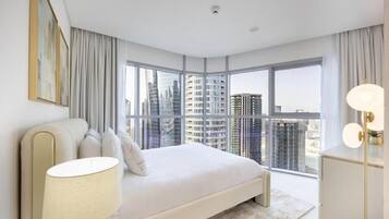 A modern bedroom with a large bed, a bedside lamp, and a view of high-rise buildings.