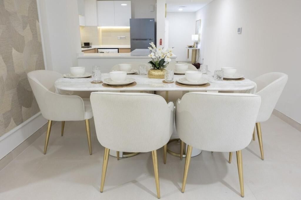 A dining area with a round table set for six, surrounded by white chairs with gold legs.