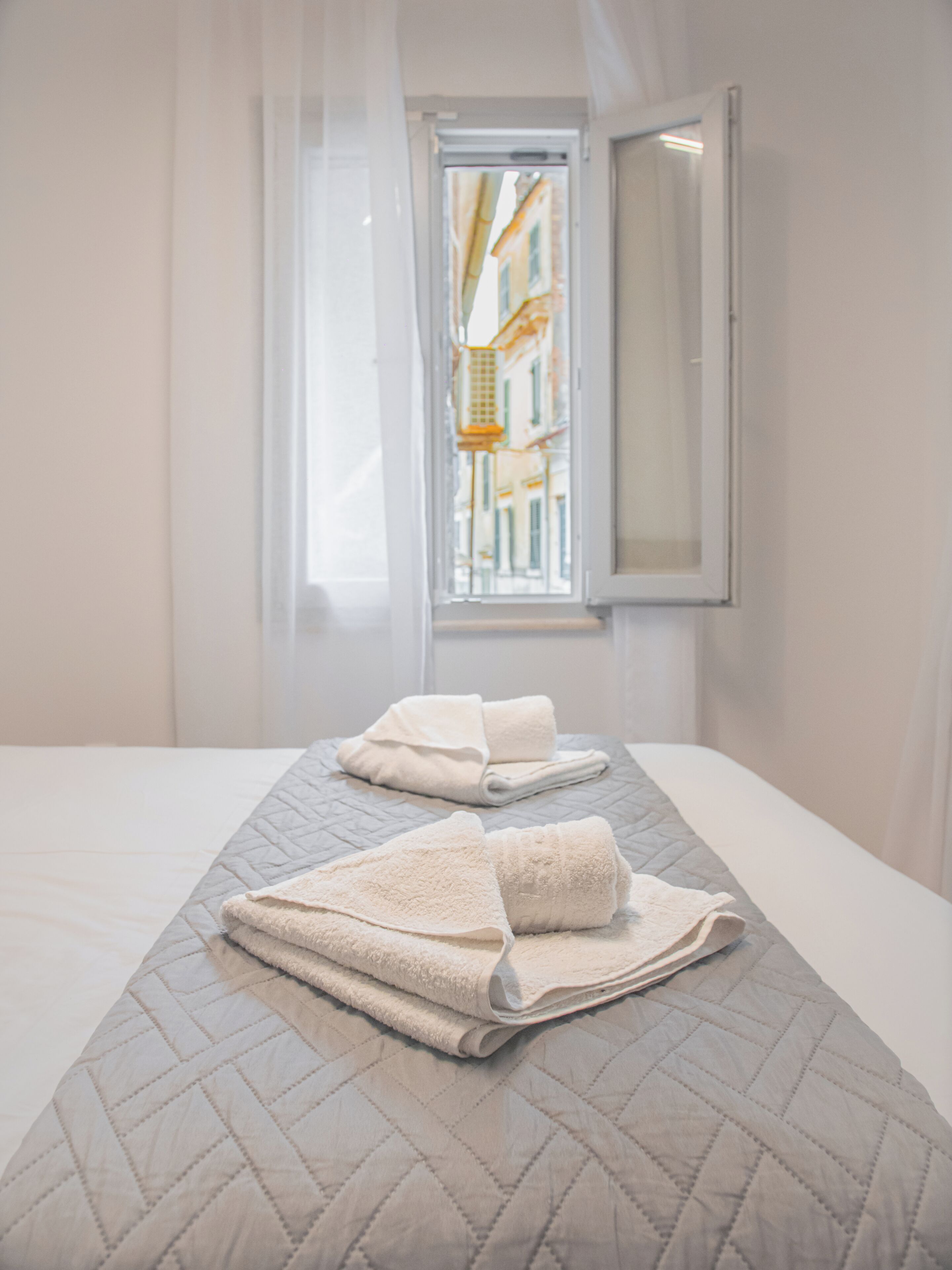 Habitación de hotel con cama, dos toallas, una ventana con cortinas y vistas a edificios.