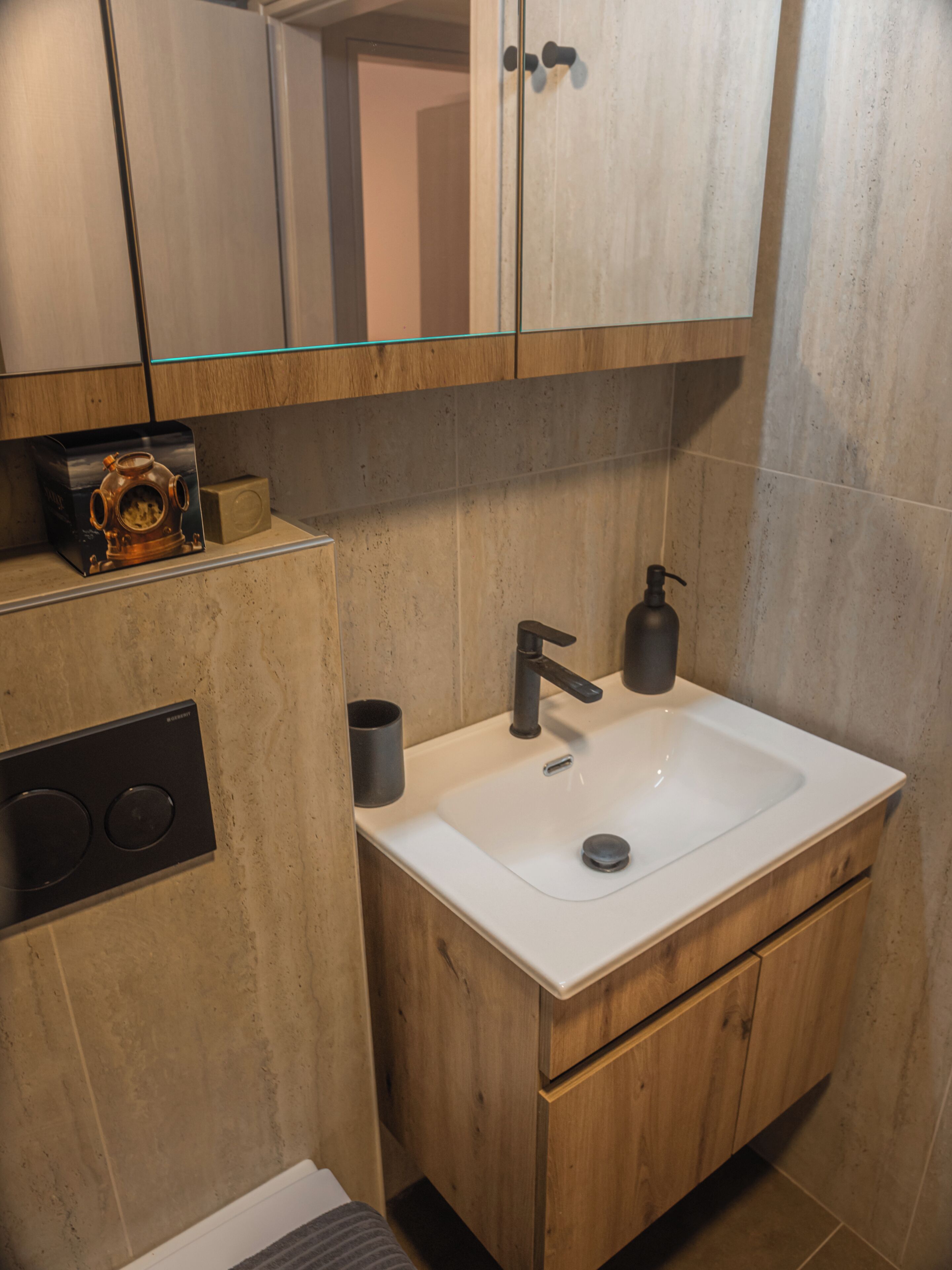 Un baño moderno con un lavamanos blanco, armarios de madera, una repisa de vidrio con un objeto decorativo y un dispensador de jabón negro.