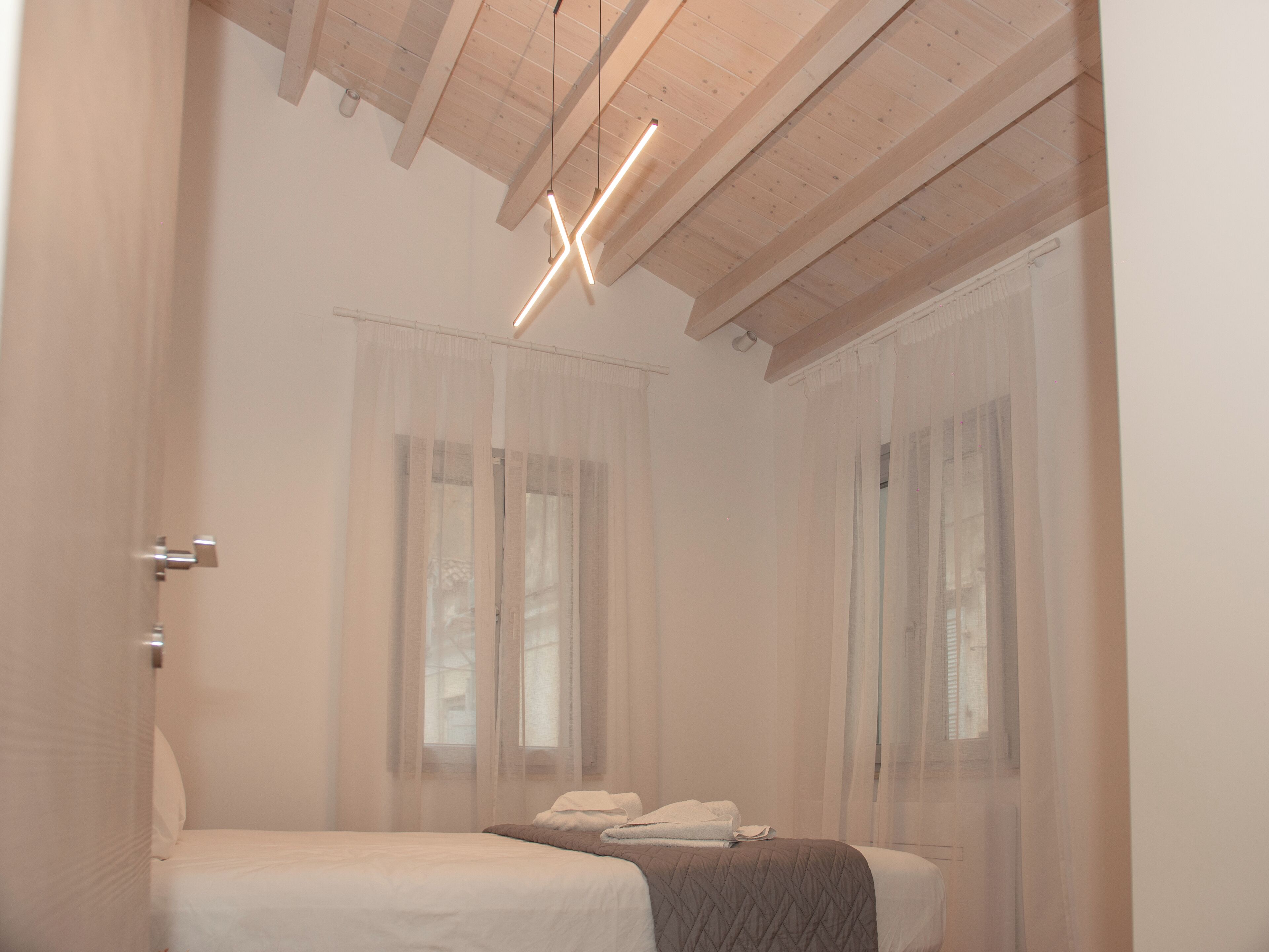 Un dormitorio con cama, ventana con cortinas translúcidas, techo de madera y lámpara moderna.
