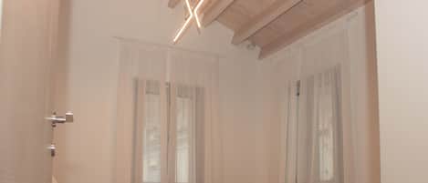 Ein Schlafzimmer mit einem Bett, einem Fenster mit durchsichtigen Vorhängen, einer Holzzieldecke und einer modernen Leuchte.