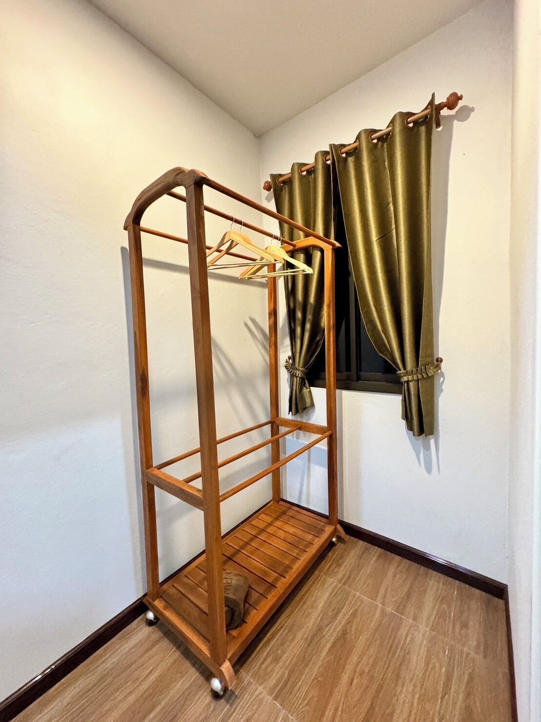A corner room with a wooden floor, a wall-mounted wooden rack with a towel, and a window with green curtains.