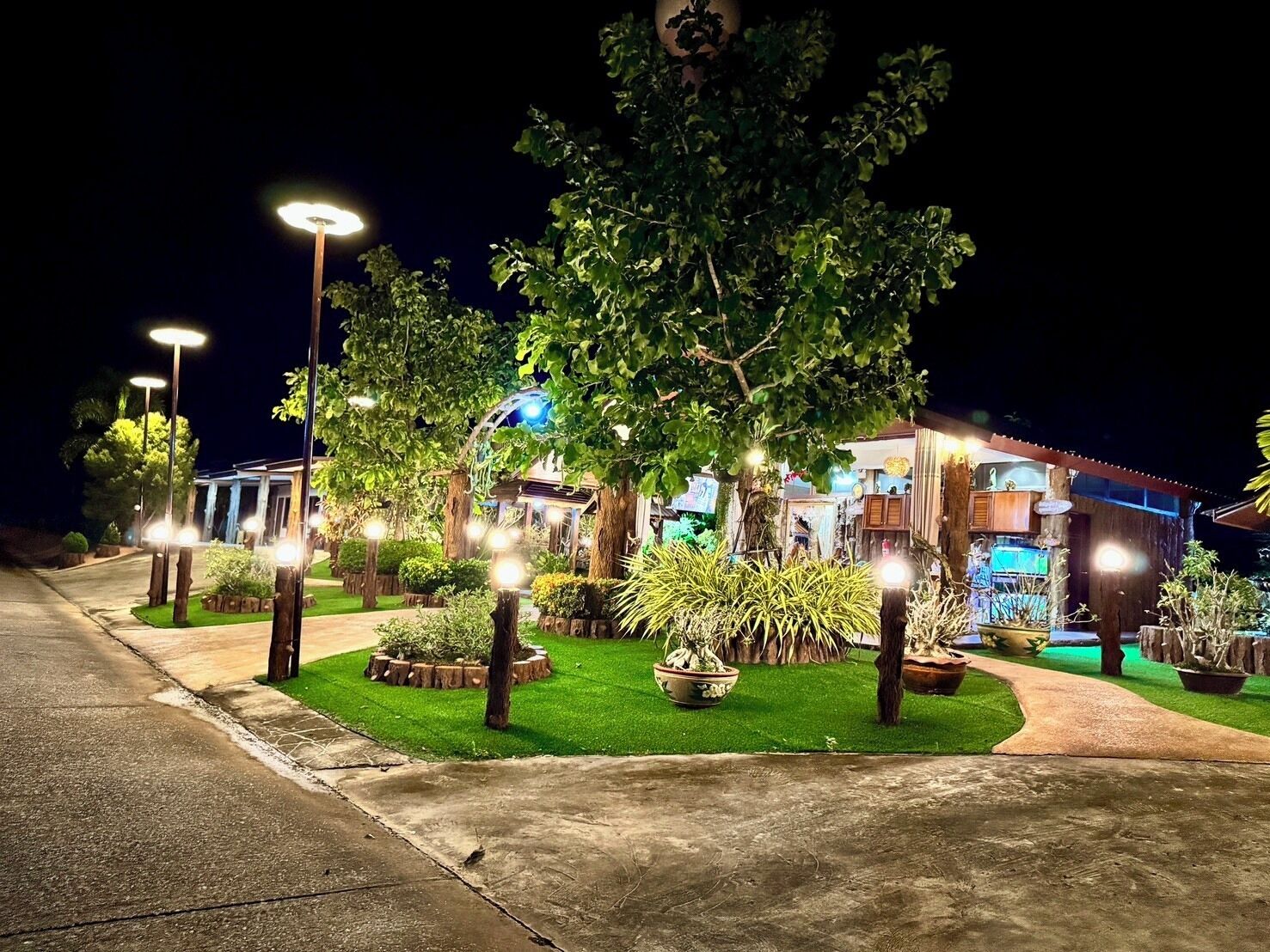 A well-lit outdoor area with a pathway leading to a building, surrounded by trees and potted plants.