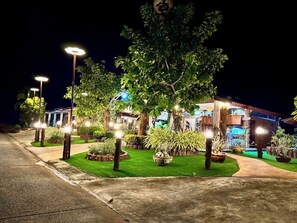 Un espacio exterior bien iluminado con un sendero que lleva a un edificio, rodeado de árboles y plantas en macetas.