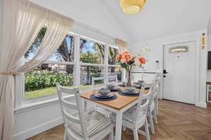 A dining area with a wooden table, white chairs, and a view of the outdoors through large windows. - The Banana Cabana - Cozy Cottage (West Palm Beach)