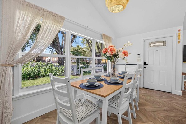 A dining area with a wooden table, white chairs, and a view of the outdoors through large windows.