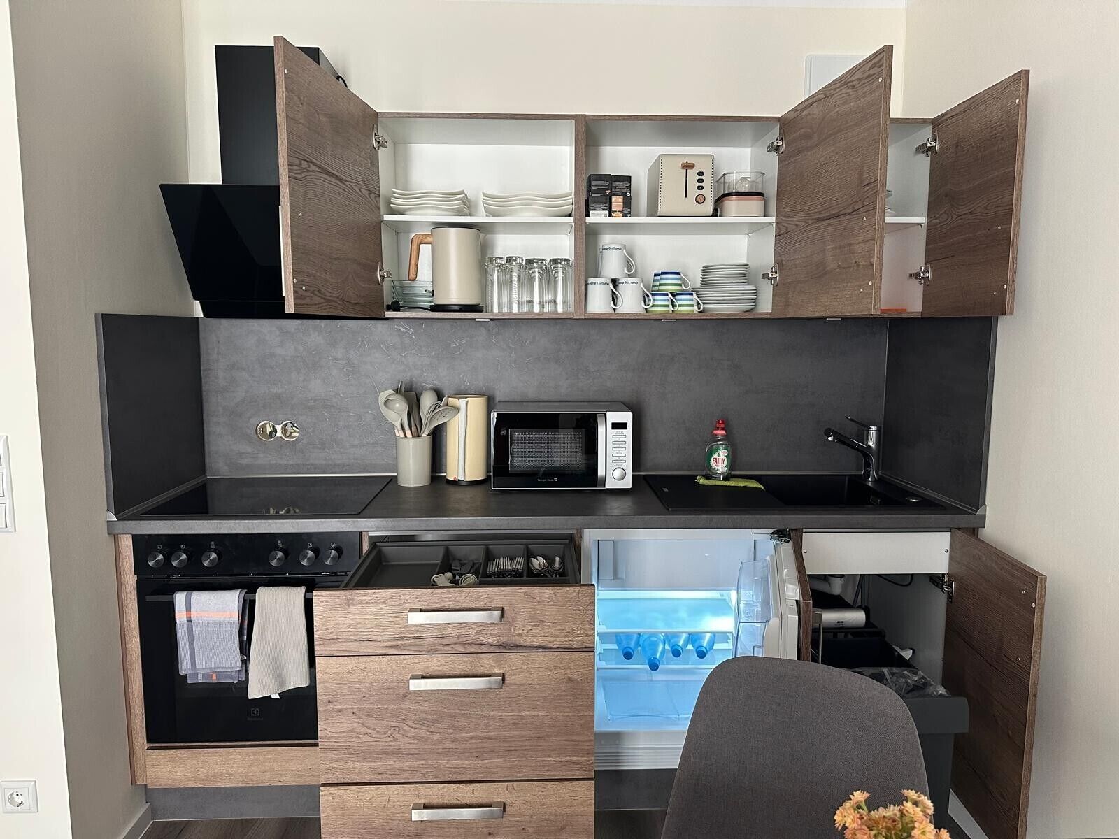 A compact kitchen with a built-in oven, microwave, and refrigerator, featuring wooden cabinetry and a black countertop.