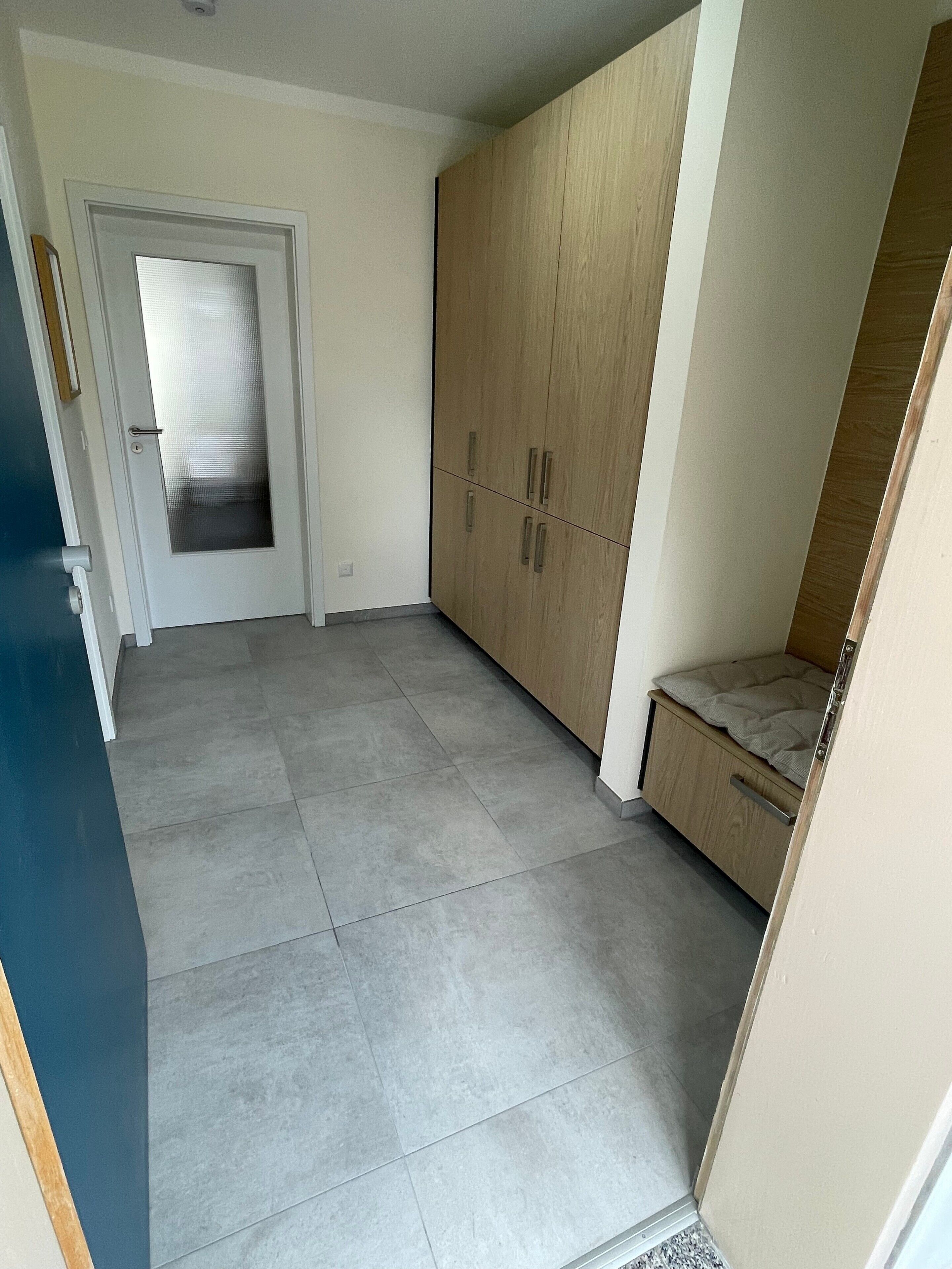 Ein Zimmer mit Fliesenboden, einem Schrank mit zwei Türen, einer Bank und einer Tür mit Milchglasfenster.