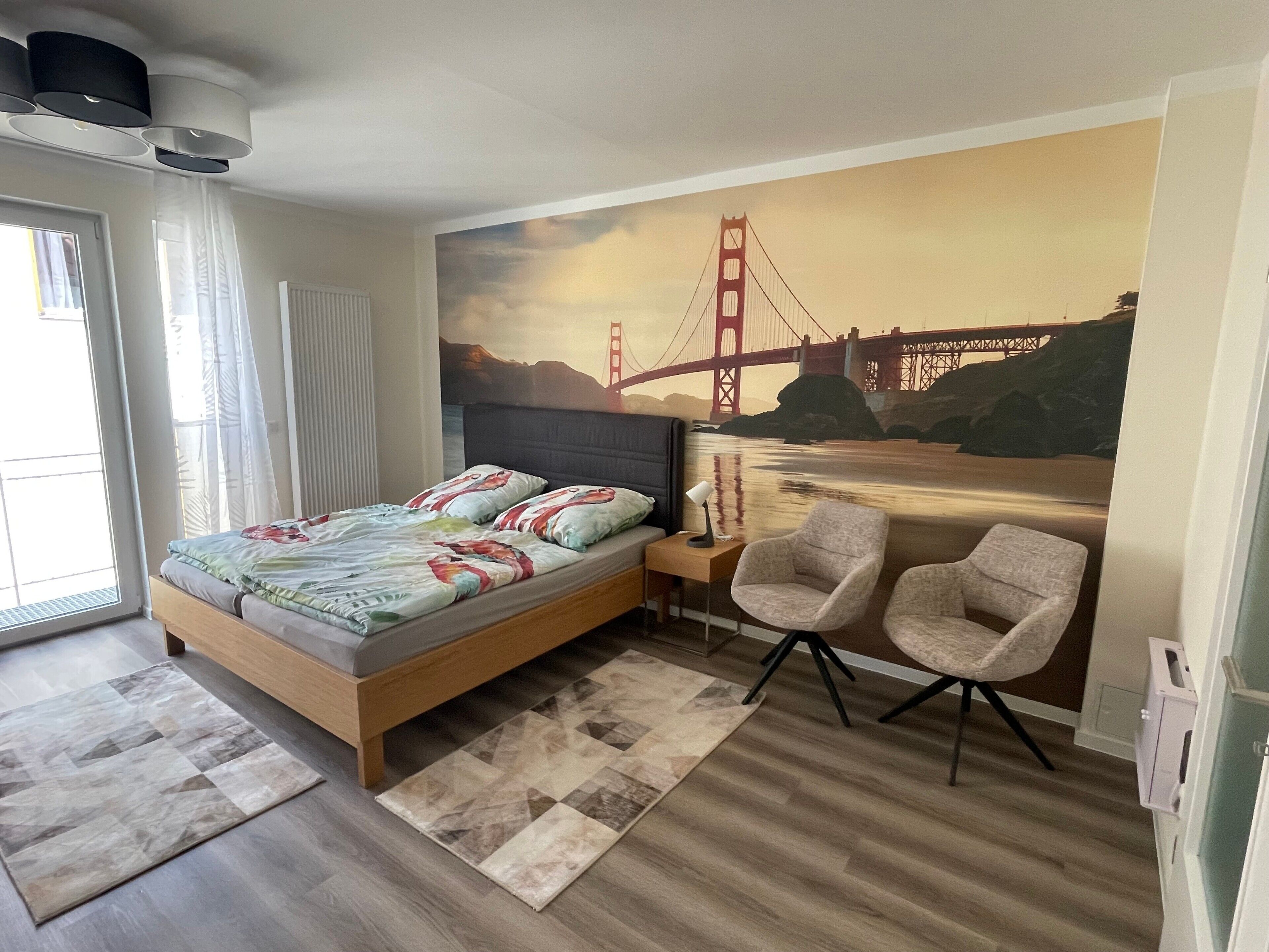 A bedroom with a bed, a nightstand, two chairs, a wall mural of the Golden Gate Bridge, and a window with curtains.