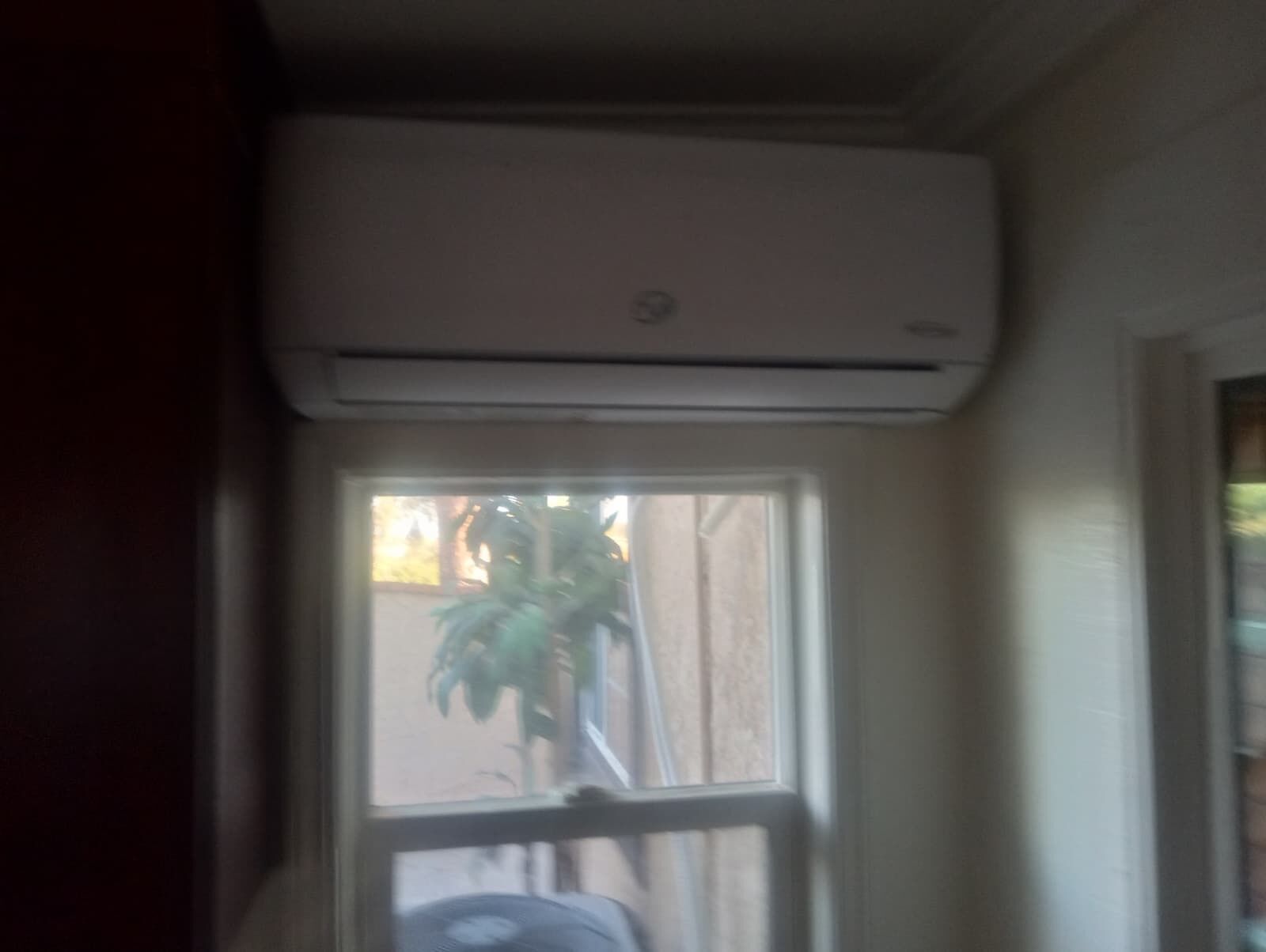 Ein Zimmer mit Fenster, einer darüber angebrachten Klimaanlage und einem Blick auf eine Pflanze vor dem Fenster.