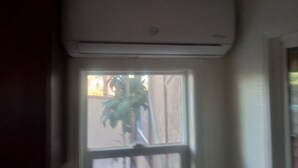 Habitació amb finestra, un aparell d'aire condicionat muntat a sobre i vista parcial d'una planta a l'exterior.