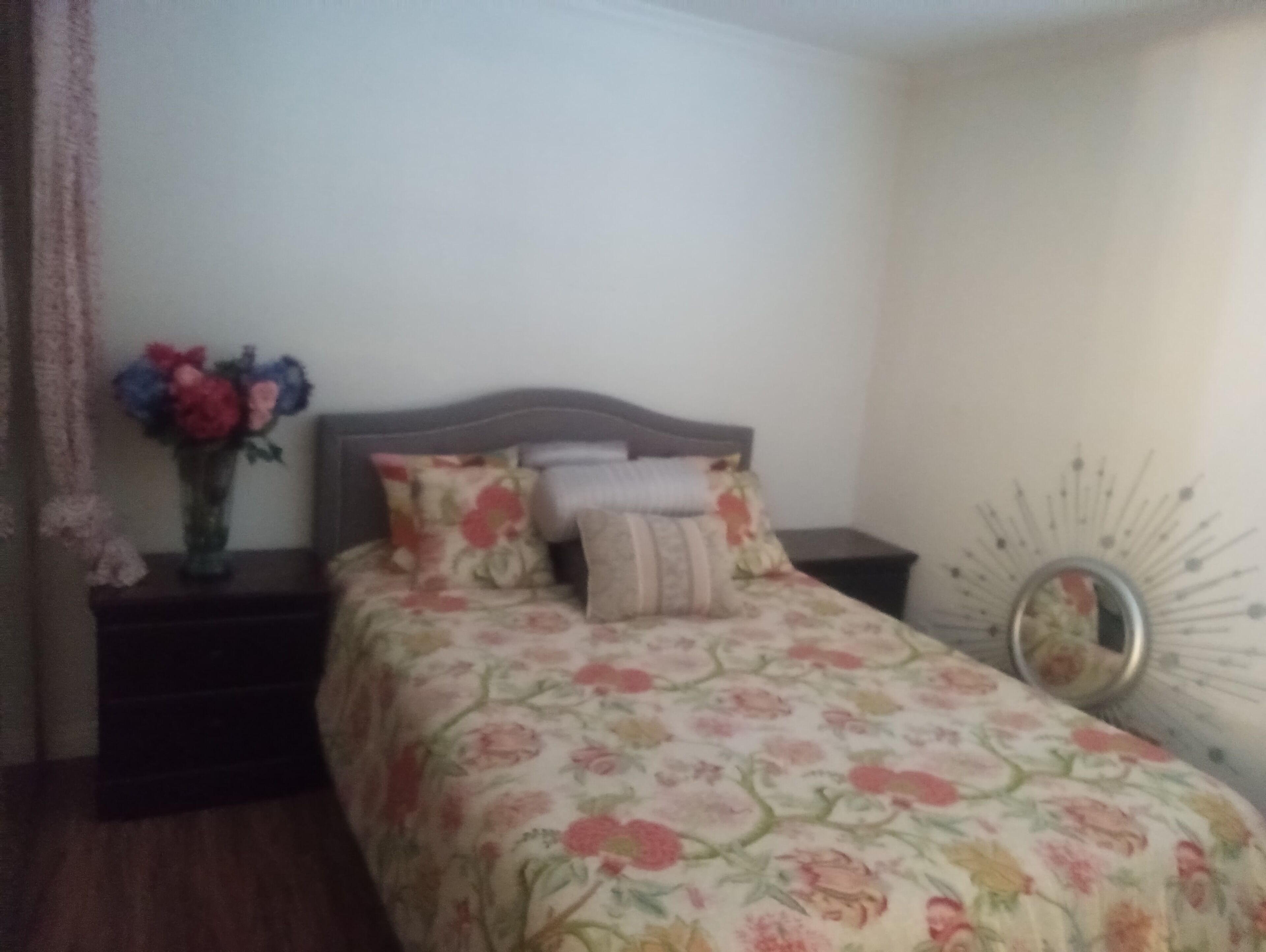 Un dormitorio con edredón estampado con motivos florales, una mesita de noche, un florero con flores y una decoración de pared.