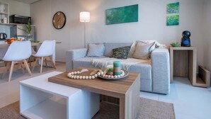 Ein modernes Wohnzimmer mit einer Couch, einem Couchtisch und Sesseln. Es gibt eine Küchenzeile mit einer Uhr und einer Kaffeemaschine.
