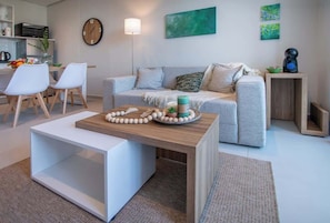 A modern living room with a sofa, coffee table, and chairs. There is a kitchen area with a clock and a coffee machine. - Corporate Stays First Living - 504 (Asunción)