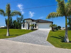 A modern two-story house with a paved driveway, palm trees, and a clear blue sky. - The Water Lily | Modern Waterfront Pool Home (Cape Coral)