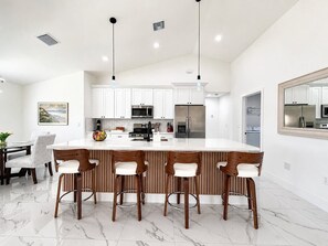 A modern kitchen with white cabinets, a central island with bar stools, stainless steel appliances, and a large mirror above the island. - Palm Paradise on the Canal (Cape Coral)
