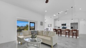 Una sala de estar moderna con un sofá, una mesa de centro de vidrio y vistas a una cocina con armarios blancos y una zona de comedor.