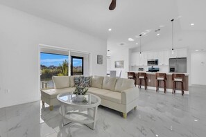 A modern living room with a sofa, a glass coffee table, and a view of a kitchen with white cabinets and a dining area. - Palm Paradise on the Canal (Cape Coral)
