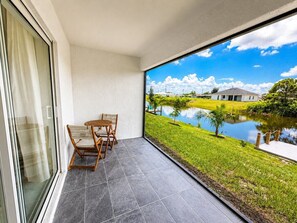 Un balcón con una mesa y sillas, vista a un lago, y un espacio verde con una casa al fondo.