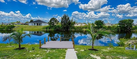 A pathway leading to a dock over a calm body of water, surrounded by greenery and a clear sky.