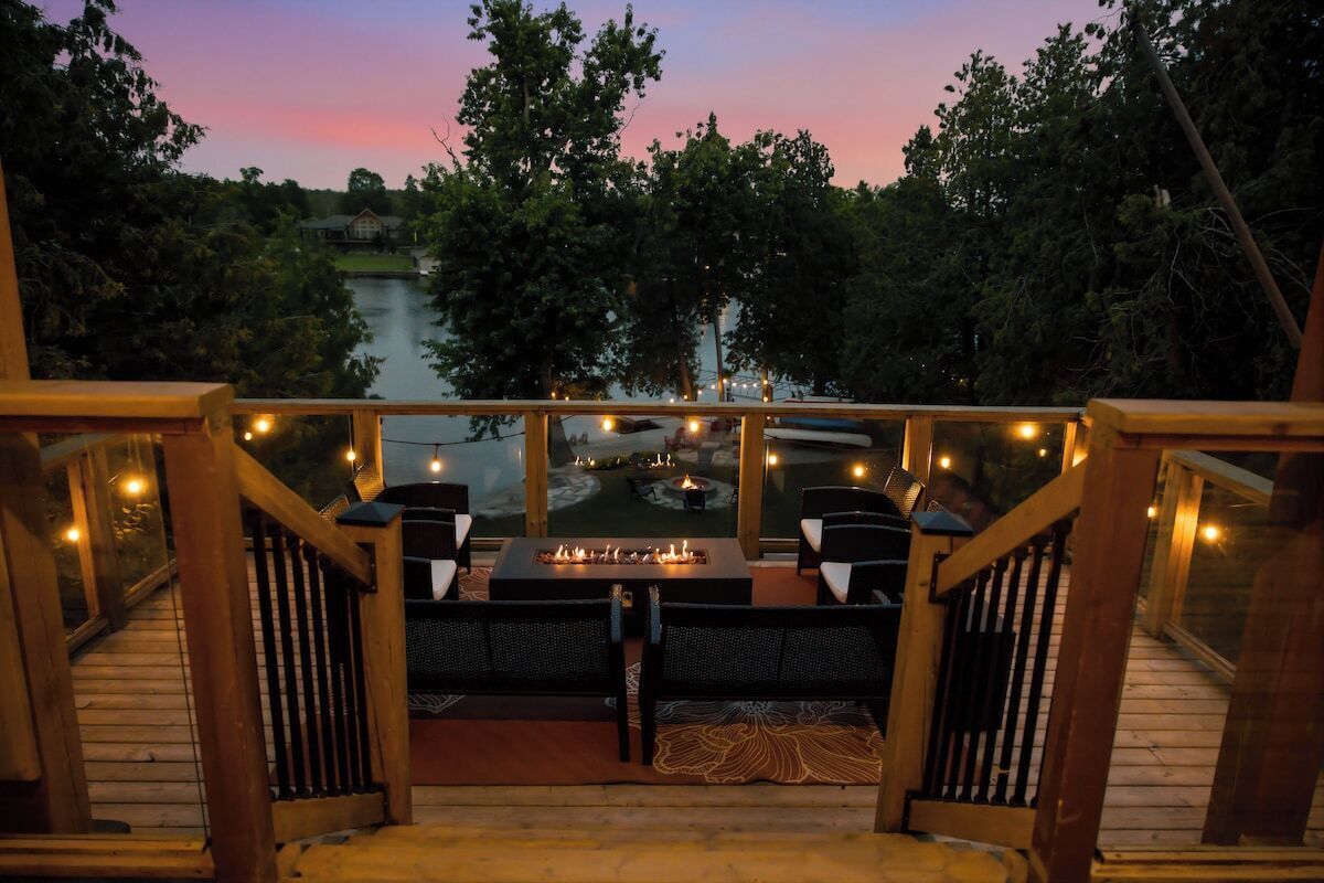 Eine Terrasse mit Sitzgelegenheiten im Freien, einer Feuerstelle und Blick auf einen See im Abendrot.