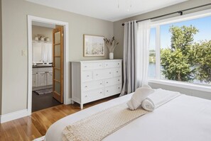 Ein Schlafzimmer mit einem großen Bett, einer Kommode, einem Fenster mit Blick auf Bäume und einem durch eine offene Tür einsehbarer Badezimmer.