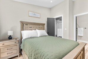 Um quarto com uma cama de madeira, uma mesa de cabeceira com abajur, uma moldura com uma frase na parede e uma porta que dá para um banheiro.