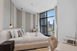 Ein modernes Hotelzimmer mit einem großen Bett, einem Schreibtisch mit einem Laptop, einem Stuhl und Blick auf die Skyline durch ein großes Fenster.