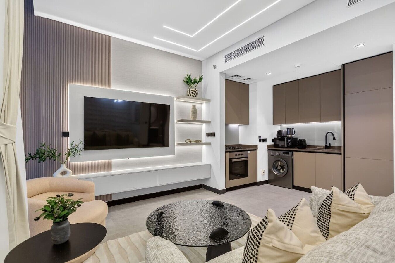A modern living room with a flat-screen TV, sofa, coffee table, and a kitchen area with appliances.