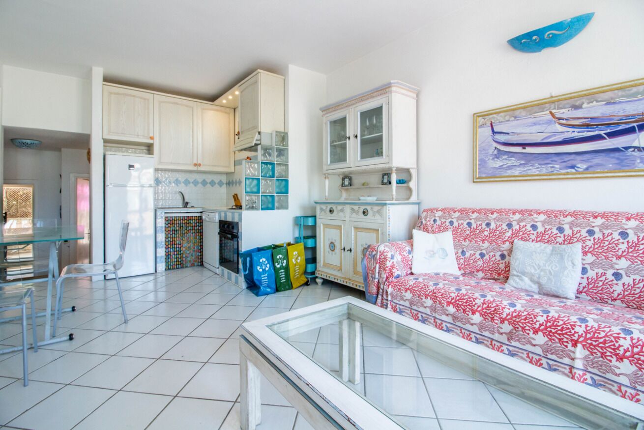 Uma sala de estar com sofá, uma mesa de centro de vidro e uma área de cozinha com armários e geladeira.