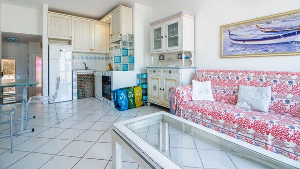 A living room with a sofa, a glass coffee table, and a kitchen area with cabinets and a refrigerator. - GuestHost - Palau Comfy Flat with Balcony (Palau)