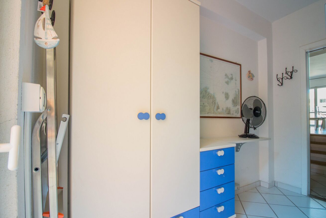 A room with a white wardrobe, blue drawers, a fan, a framed picture, and a door with a handle.
