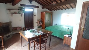 Una stanza accogliente con un tavolo di legno, sedie, un camino, un letto con copriletto verde e una porta che dà su un'altra stanza.