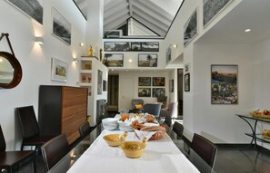 A dining area with a long table set for a meal, surrounded by chairs, with artwork on the walls and a high ceiling with skylights. - Cirasa Luxury Country House (Trecastagni)