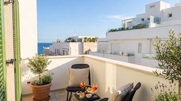 A balcony with a potted plant, a chair, a small table with food and drinks, and a view of the sea and buildings.