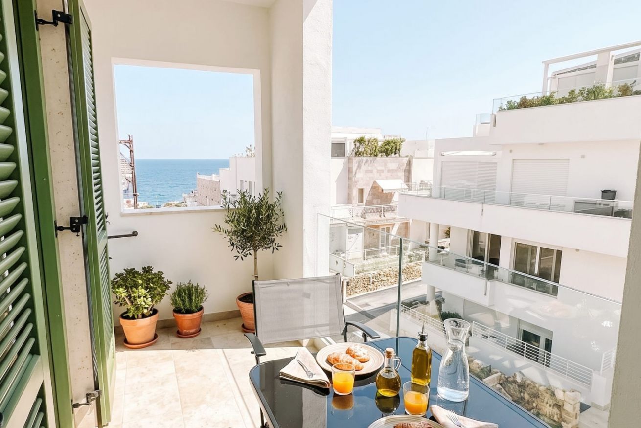 A balcony with a table set for breakfast, potted plants, and a view of the sea and buildings.