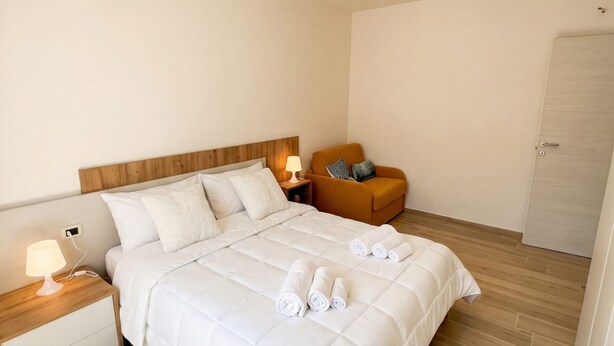 Ein ordentlich gemachtes Bett mit weißer Bettwäsche und gerollten Handtüchern, ein Holz-Kopfteil, ein Nachttisch mit Lampe und ein brauner Sessel mit Kissen.