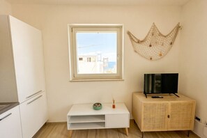 Ein Zimmer mit einem weißen Schrank, einem Fernseher darauf, einem kleinen Tisch mit Schüssel und Vase sowie einem Fenster mit Blick auf Gebäude.