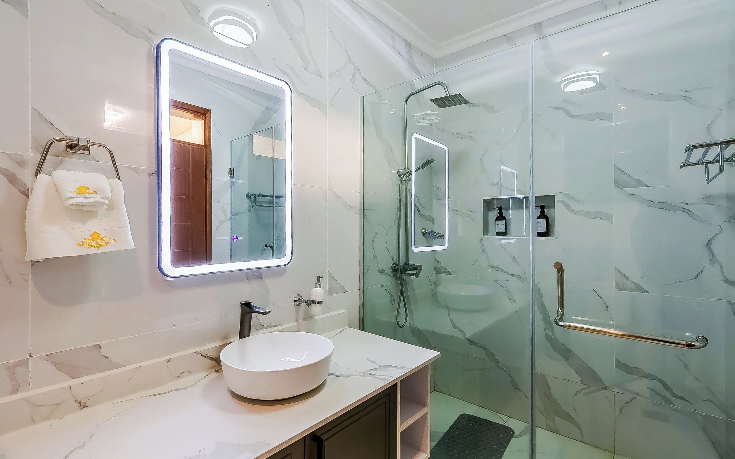 Un bagno con un vanità in marmo, un lavandino rotondo, un box doccia in vetro, un soffione a mano, un portasciugamani con un asciugamano e uno specchio con illuminazione.