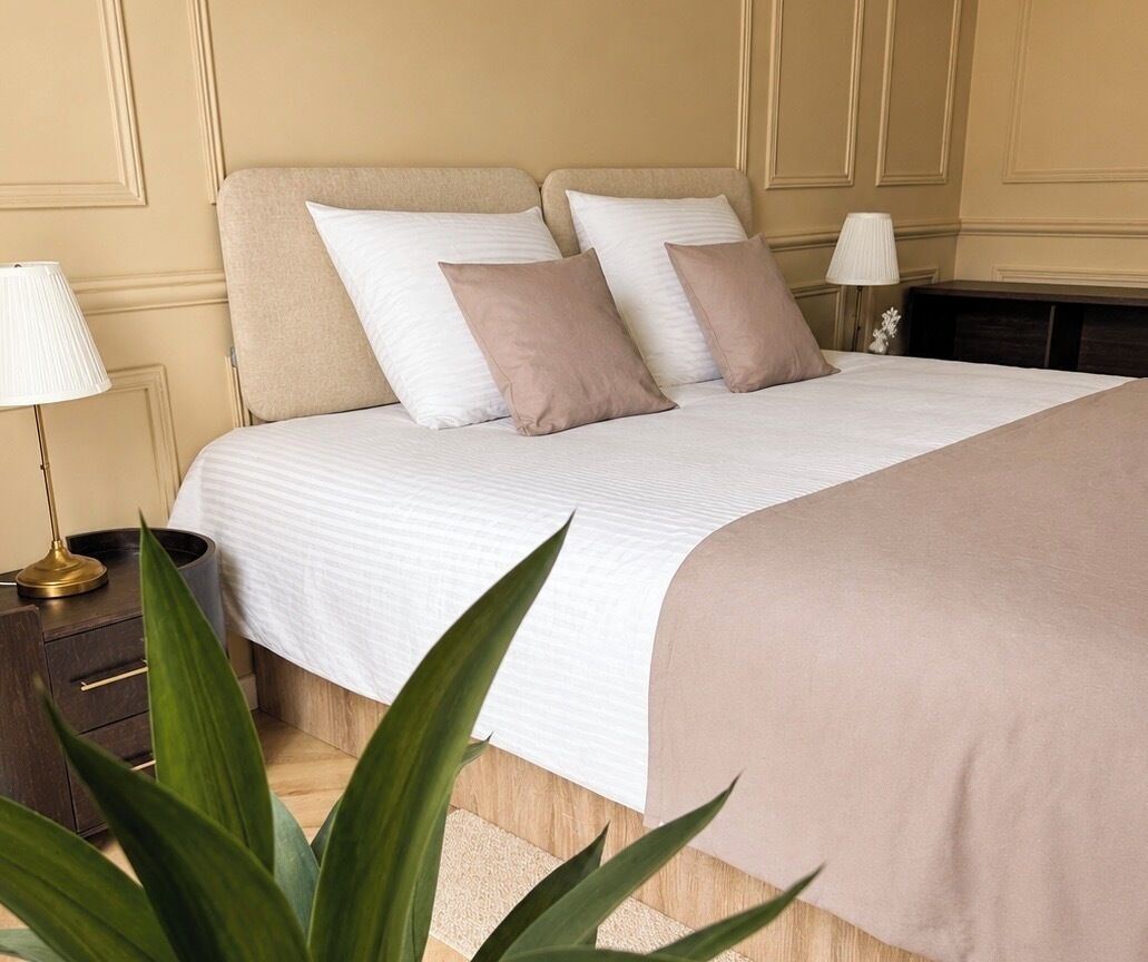 Een netjes opgemaakt bed met een wit en beige sprei, met aan weerszijden bedlampen, in een kamer met een houten vloer en een donkere nachtkastje.
