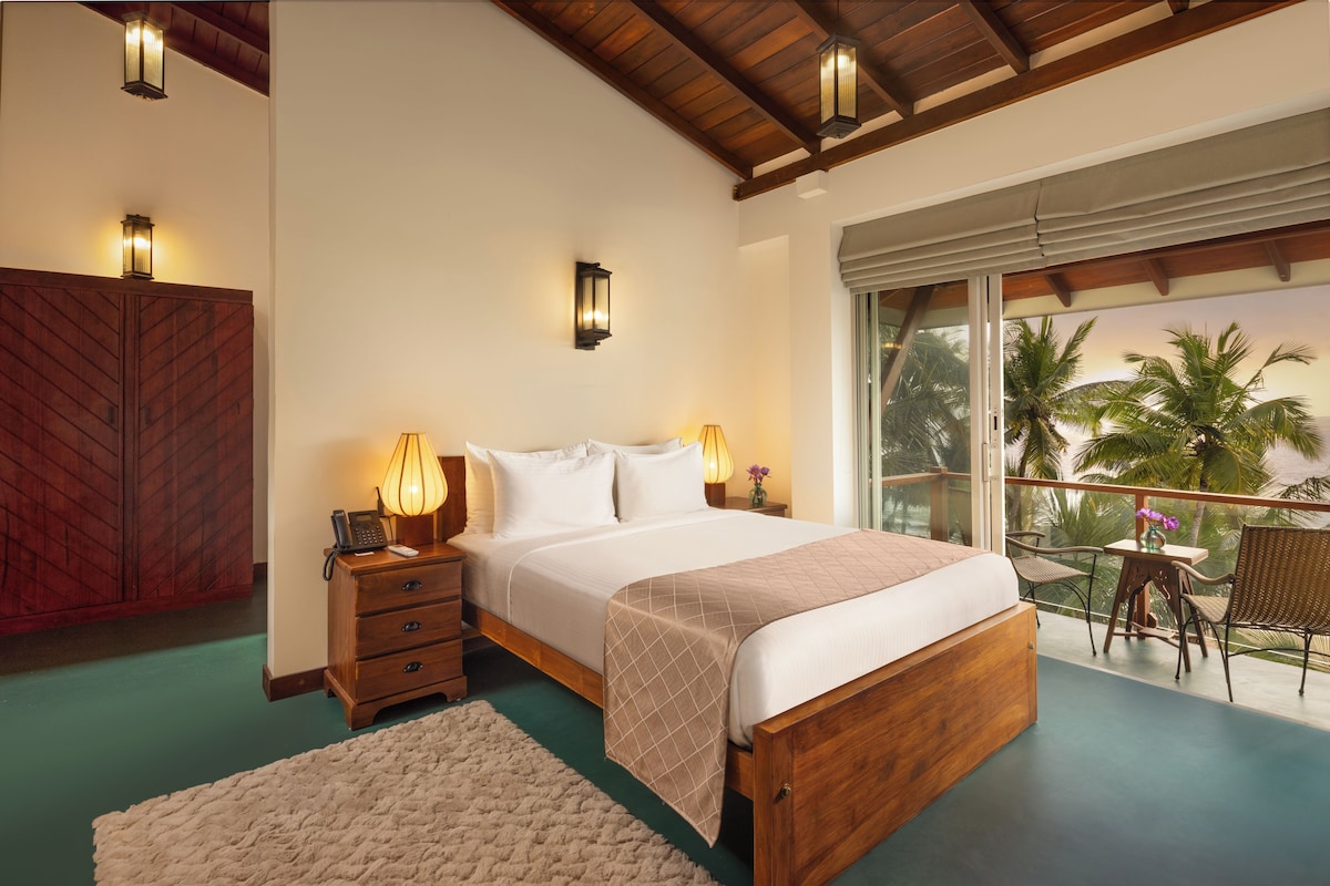 A hotel room with a bed, bedside lamps, a wooden wardrobe, a small table with a vase, and a balcony with a view of palm trees.