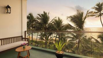 A balcony with a view of the beach and palm trees, a wooden bench, a small table with a vase, and a potted plant.
